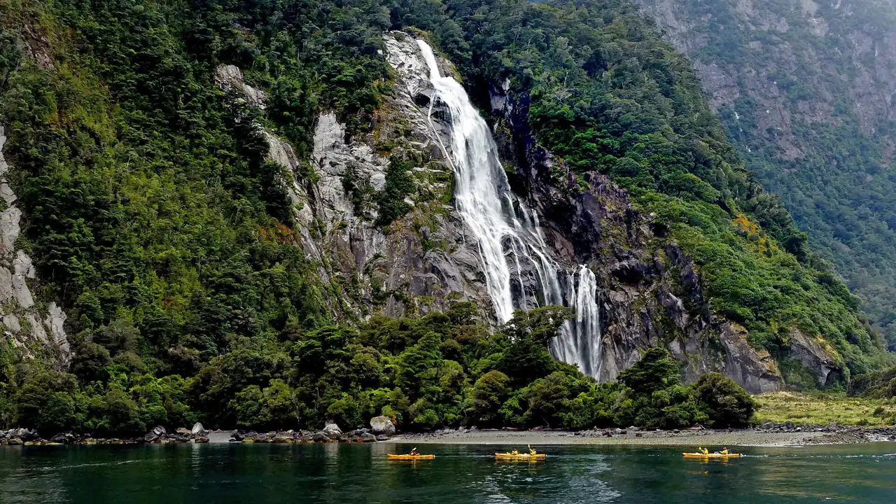 Ein Wasserfall stürzt von einer bewaldeten Felswand in den Fjord, unten kleine Kajaks auf dem Wasser