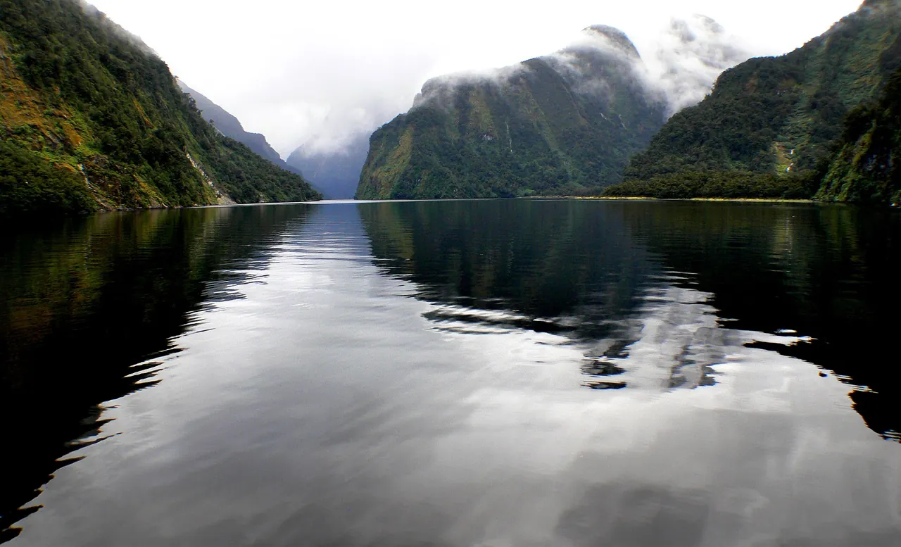 Doubtful Sound con paredes boscosas reflejadas en el agua en calma y nubes en las crestas